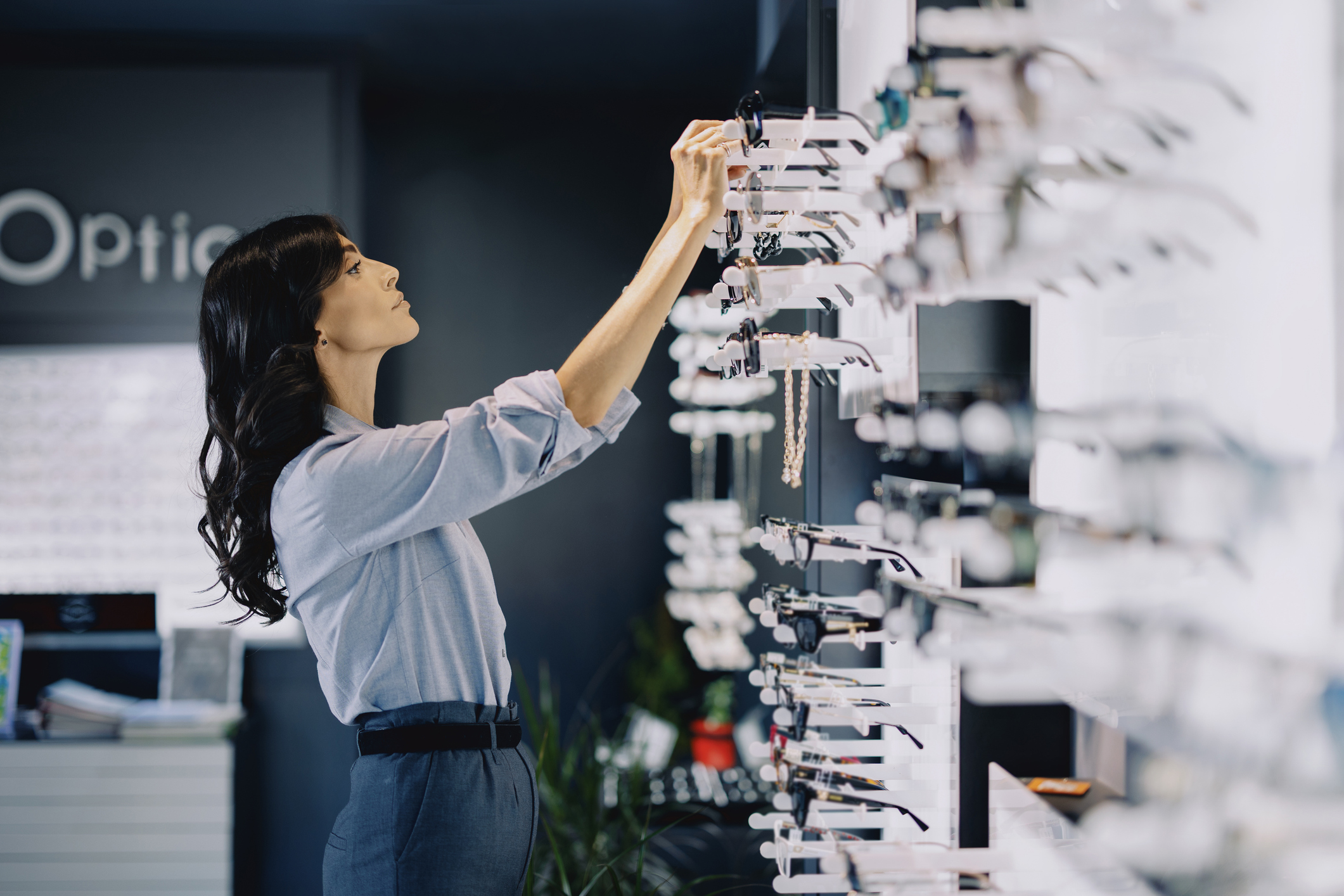 Attractive young woman trying on eyeglasses in a modern optical store. She is standing by a display shelf, thoughtfully examining different frames. The store is bright and stylish, with a wide selection of eyewear visible in the background
