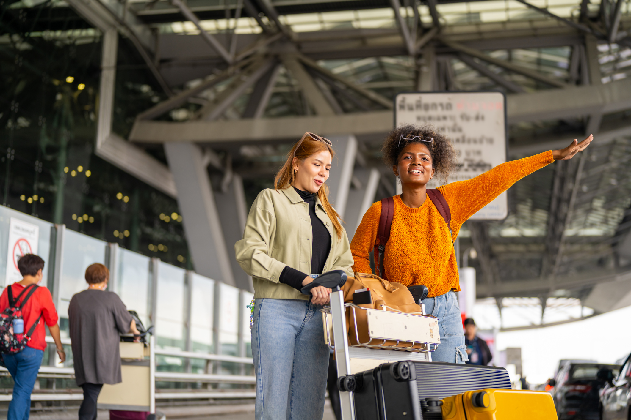 Happy woman friends with luggage trolley waiting for taxi go to hotel in the city together at airport terminal. Attractive girl enjoy travel on holiday vacation with airplane and public transportation.