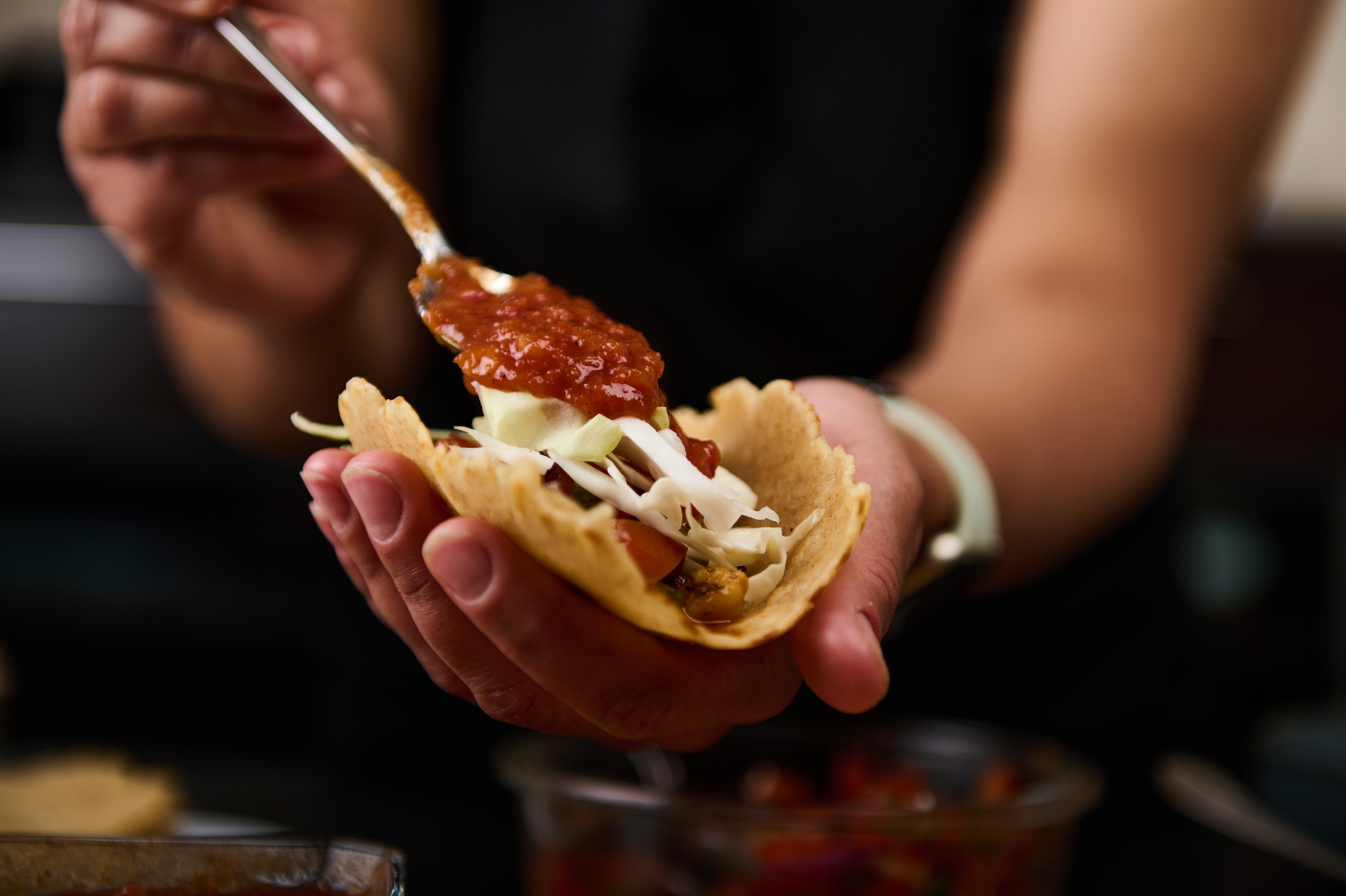 A person assembles tacos filled with fresh vegetables and vibrant salsa, showcasing delicious homemade cooking. The scene highlights culinary craftsmanship and the enjoyment of making meals.