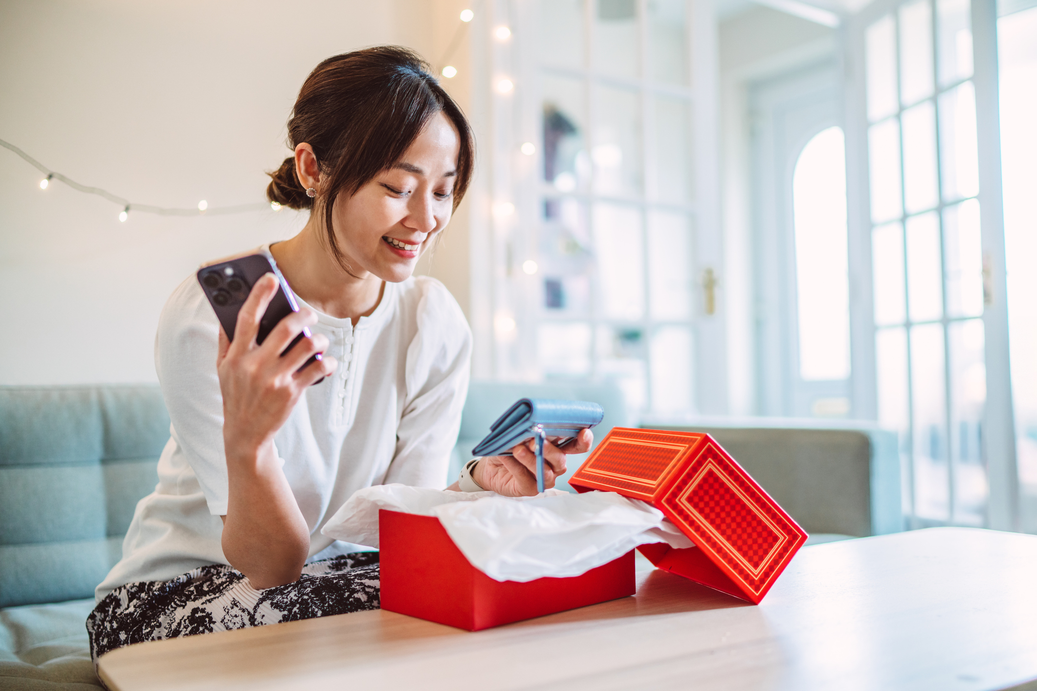Cheerful woman enjoying the excitement of unboxing her new wallet with a cheerful expression while using her smart phone to confirm her online purchase. Concepts of mobile shopping, digital lifestyle, contactless payment, and consumer behaviour.