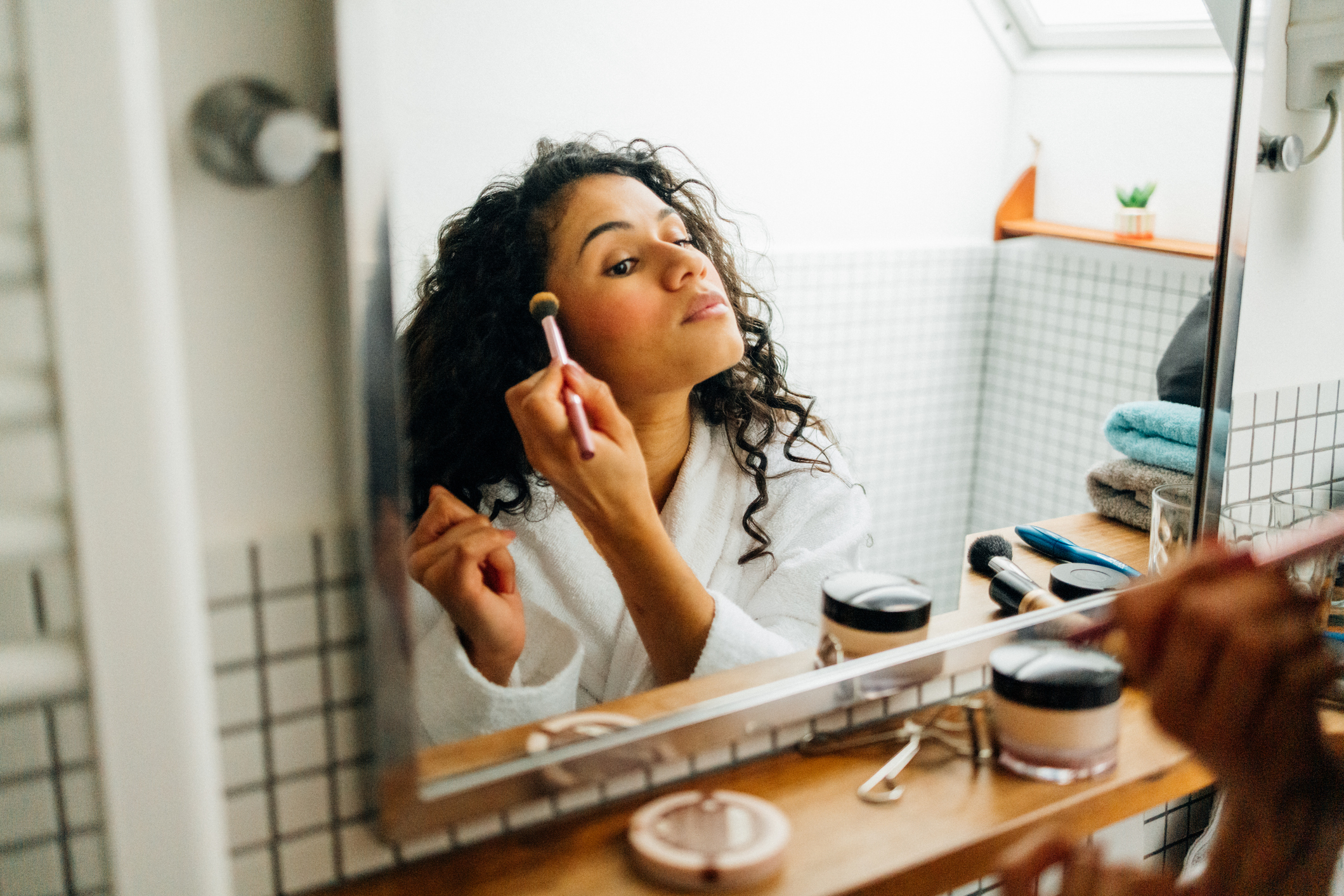 A woman in a white robe applies makeup in a bright bathroom, reflected in the mirror. She blends powder with a brush, surrounded by cosmetics, towels, and a cozy scene. Part of a series with video
