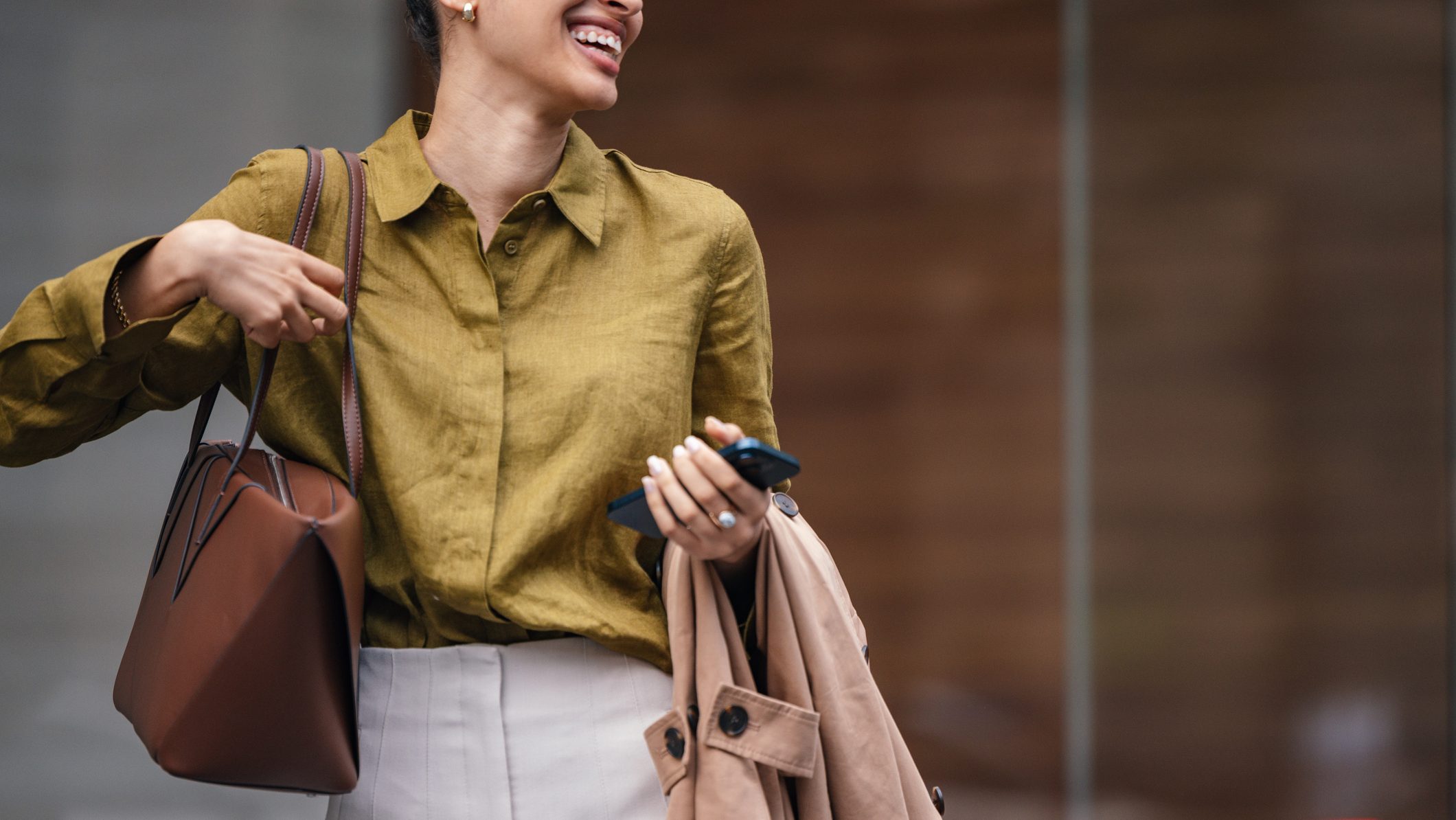 A stylish businesswoman walks outdoors in an urban setting, carrying a brown handbag and a beige coat. She smiles, exuding confidence and a positive, professional vibe as she moves through the street.
