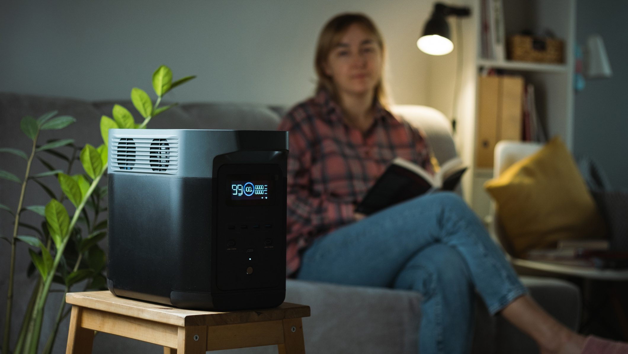 Young woman reading a book on a sofa and using Portable power station to power up lamp during electricity outage. Portable battery as a source of electricity during blackout