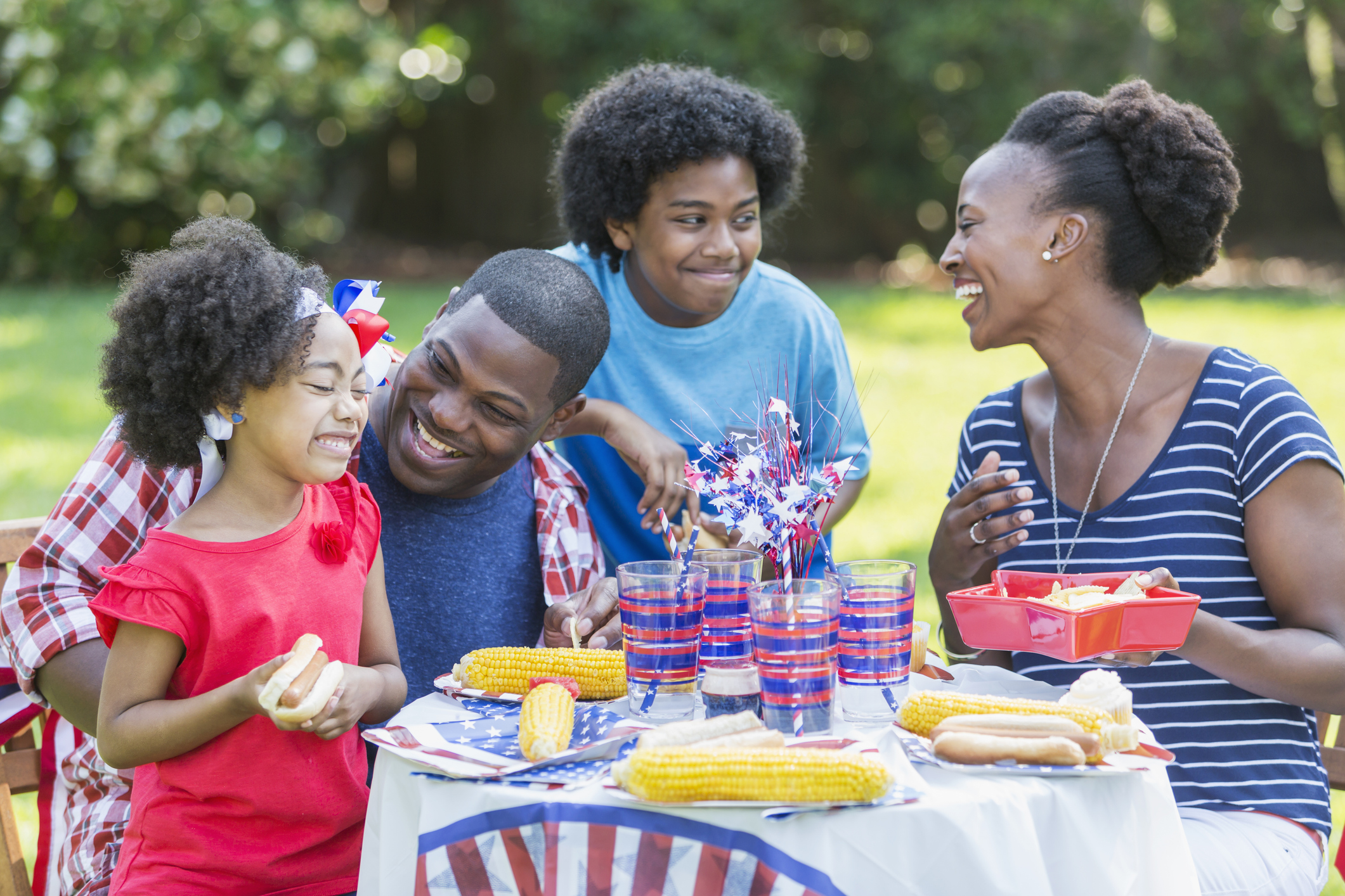 A family enjoying a backyard cookout on the 4th or July or Memorial Day. The children are mixed race African American, Asian and Hispanic. They are eating hotdogs and corn on the cob. The 12 year old boy is standing between his parents, looking at his mother.
