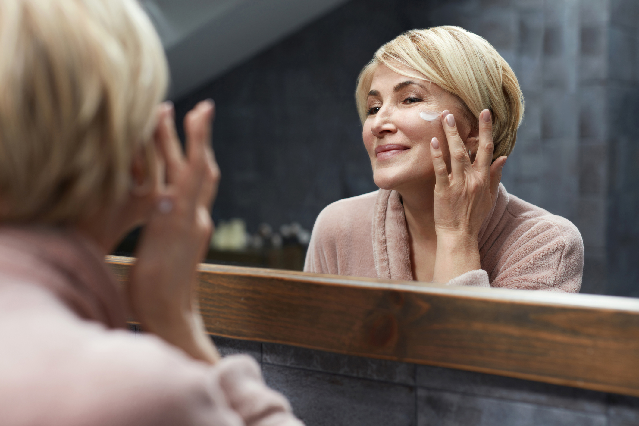 Skincare Routine. Mature Woman Uses Cosmetic Cream On Face Skin In Front Of The Mirror Reflection. Anti Aging Beauty Treatment Concept.