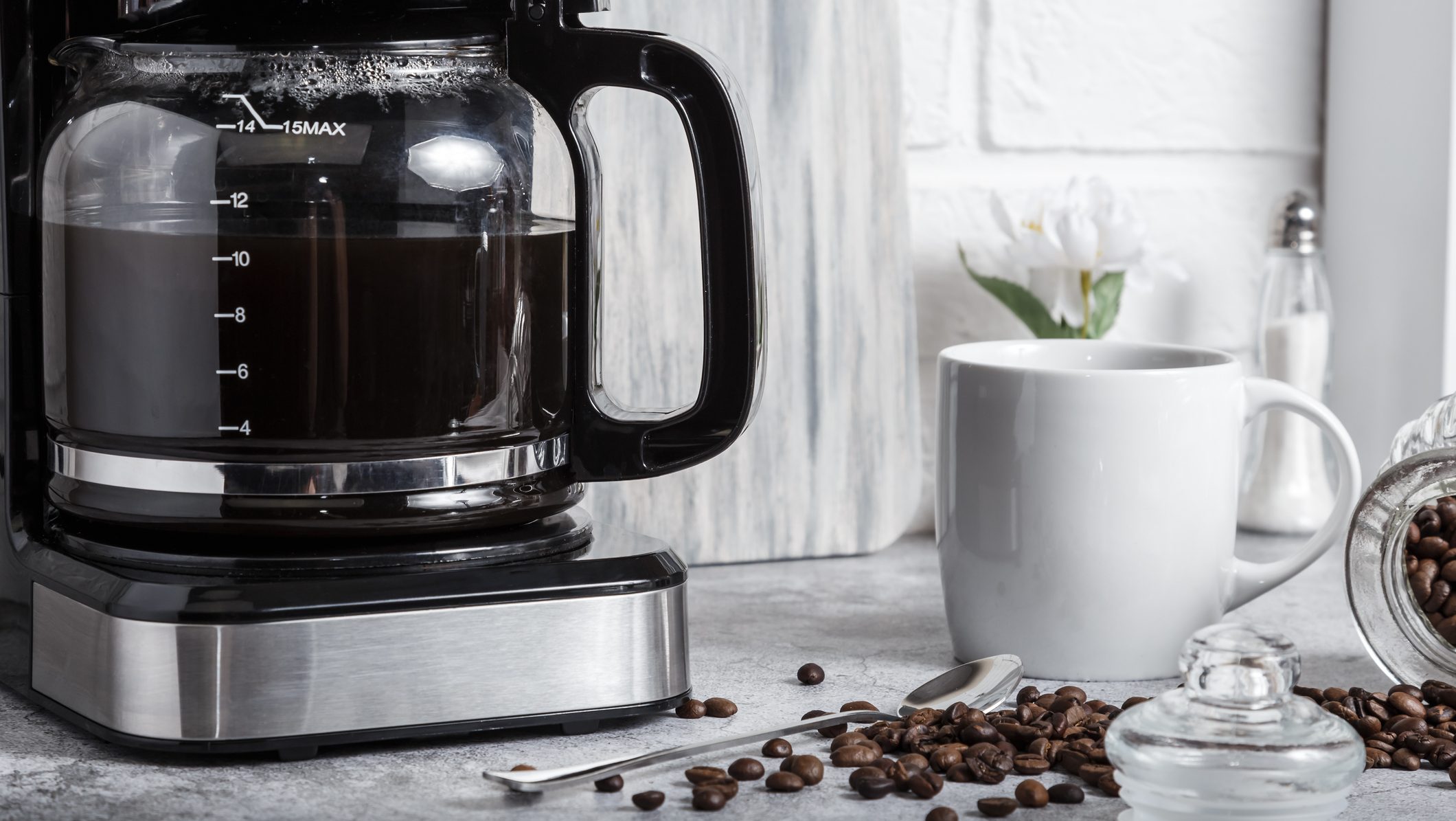 A black drip electric coffee machine with a glass teapot brews a morning drink. Household appliances, a white cup and a jar of beans on the kitchen table on the countertop at home.