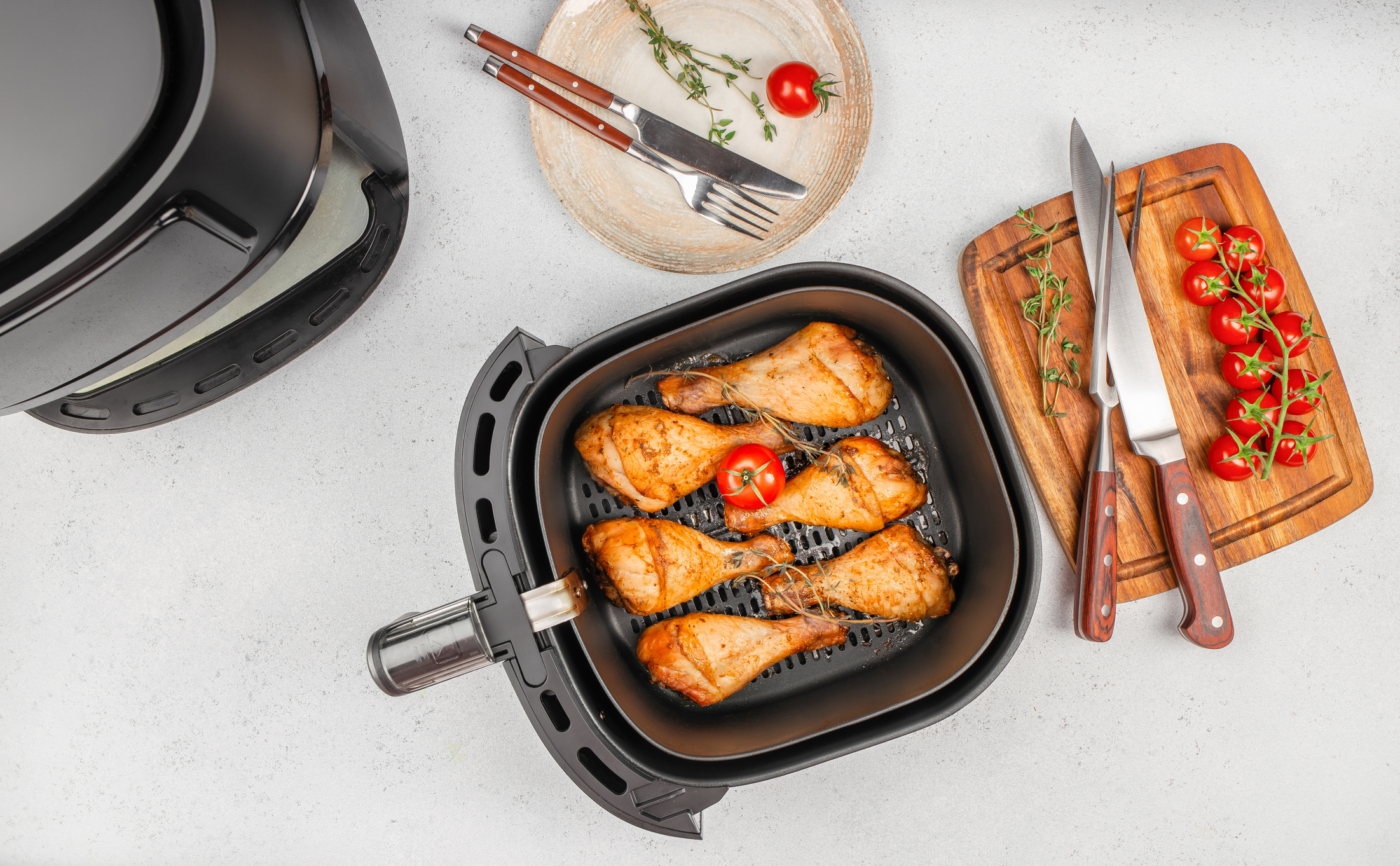 Grilled chicken drumsticks in air fryer basket with tomatoes, herbs and kitchen tools on countertop. top view. copy space