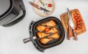 Grilled chicken drumsticks in air fryer basket with tomatoes, herbs and kitchen tools on countertop. top view. copy space