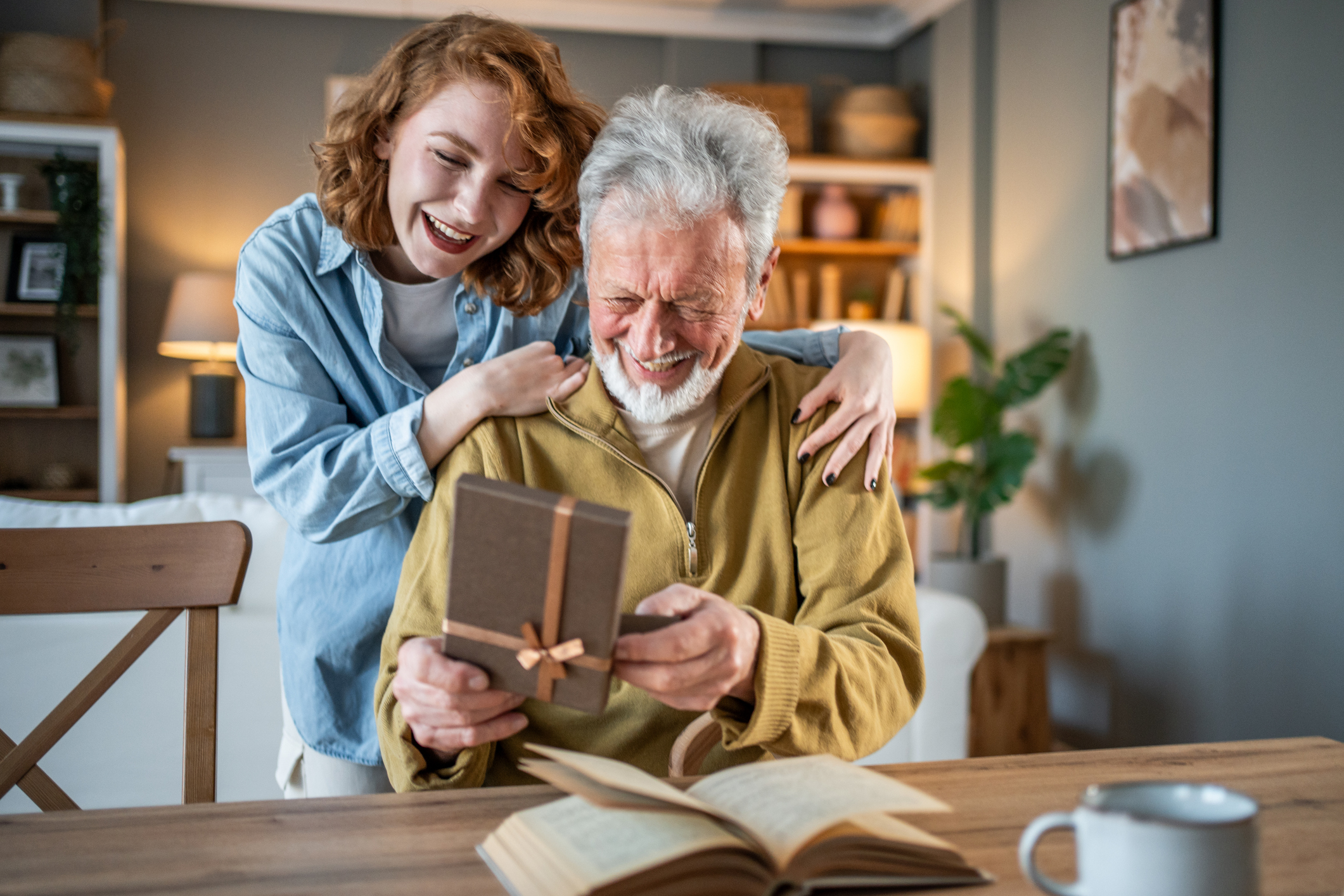 Loving granddaughter is embracing her grandfather and giving him a gift while he's sitting at the table, reading a book, in a cozy home interior