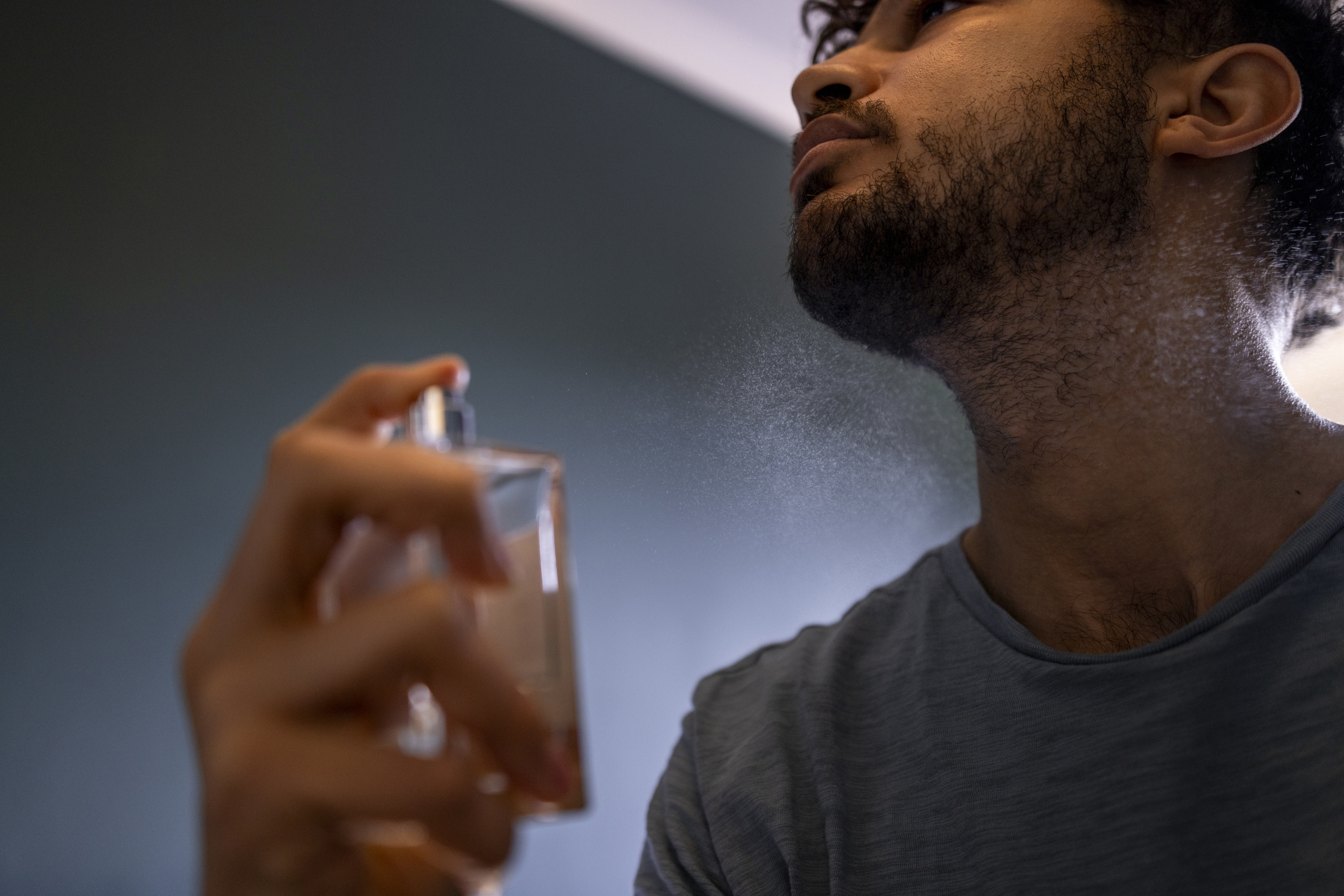 A low angle close up view of a mid adult male spraying his fragrance in his bedroom at home in the North East of England. He is spraying his favourite fragrance on his neck and looking content as he holds his hand up to his face.