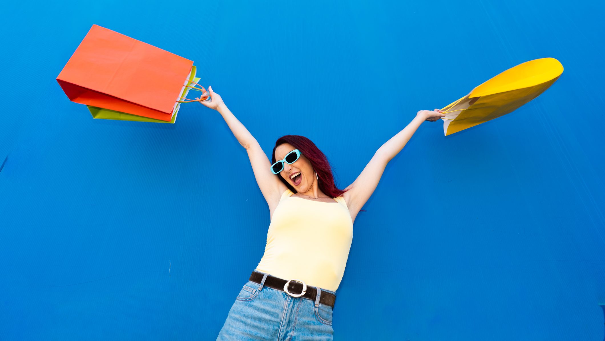 Woman standing against a blue wall, arms raised with colorful shopping bags