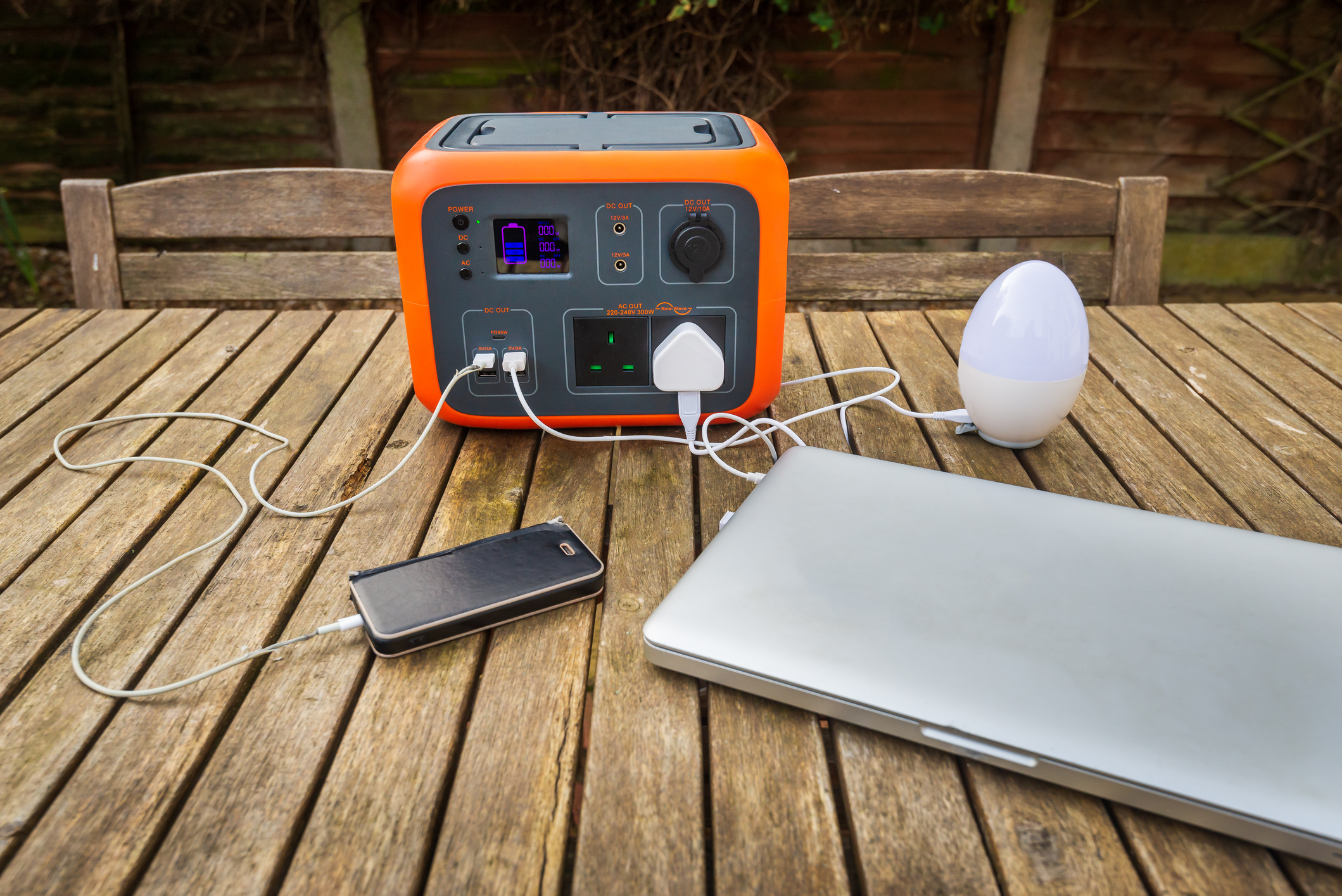 Portable power station solar electricity generator outdoors on wooden table with laptop, mobile phone and light electronic devices charging. Wireless charging lithium battery backup for use off grid.