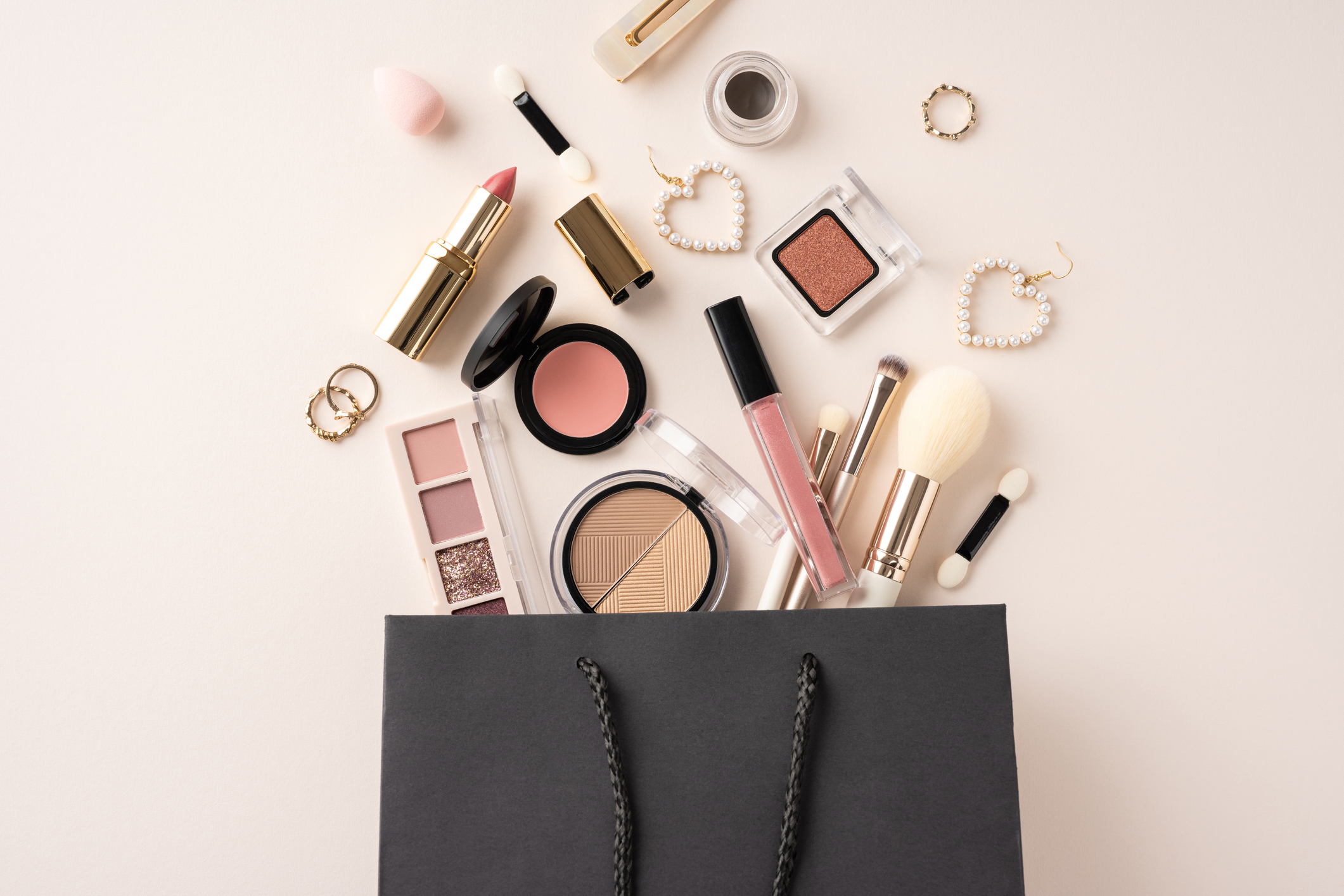 Assorted cosmetics and makeup items pouring from a black shopping bag, including lipsticks, brushes, and eyeshadow