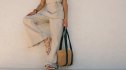 A woman poses in neutral-toned linen clothing, leaning against a textured wall, holding a woven tote bag.