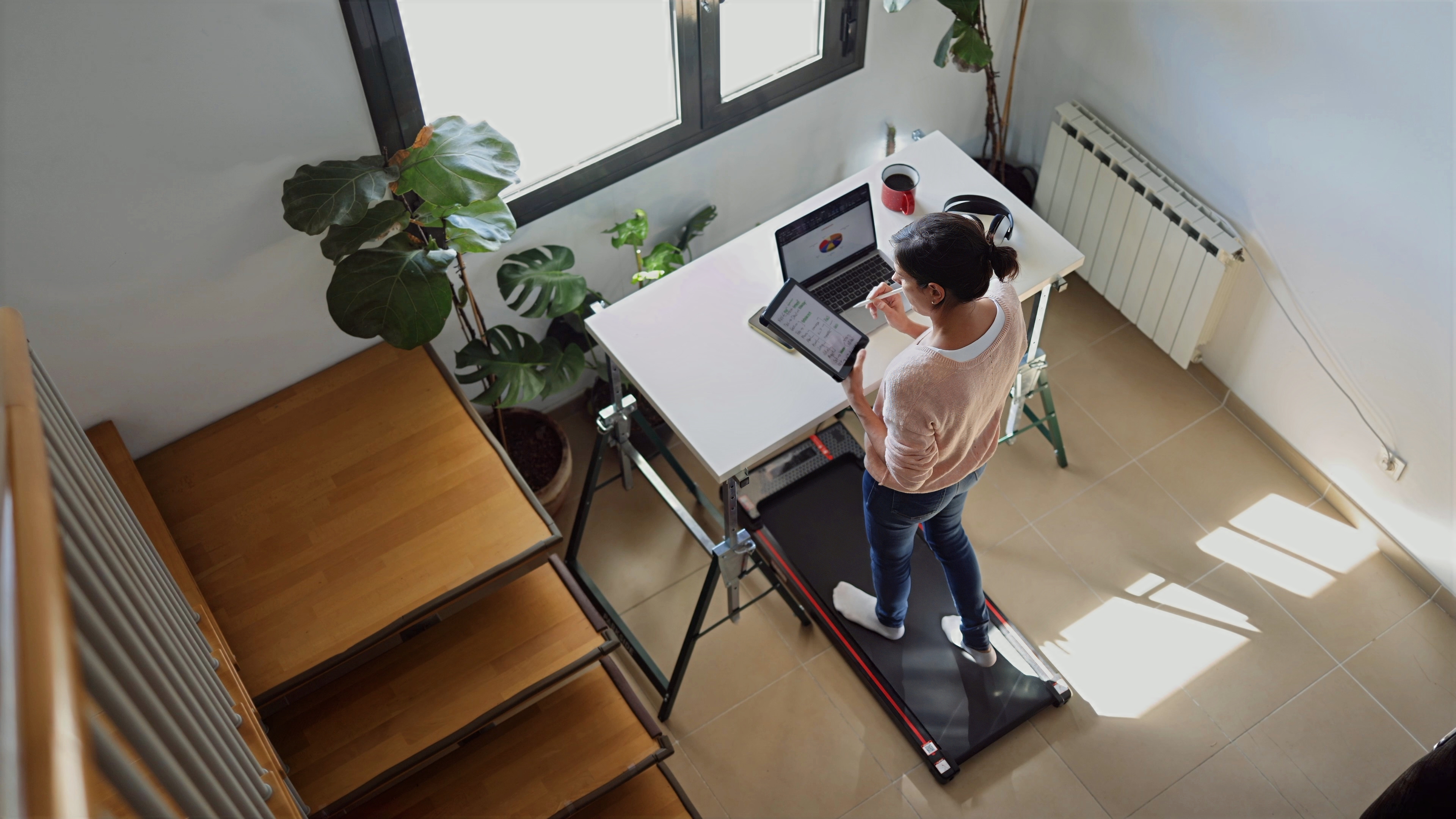 Young adult woman staying active while working remotely, walking on a treadmill desk and using a laptop and digital tablet for enhanced productivity and wellness from her home office