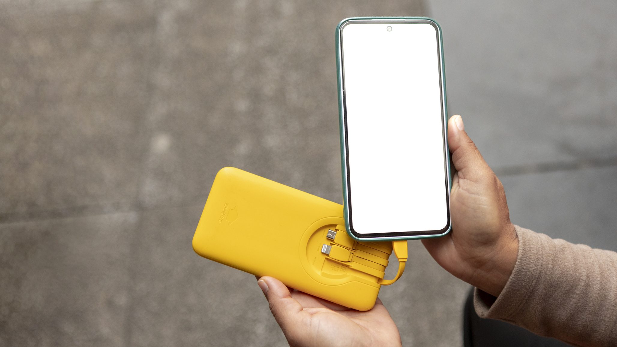 Woman holding a yellow power bank charging a smartphone with a blank white screen while traveling in the city, hands close-up conveying portable power and connectivity on the go