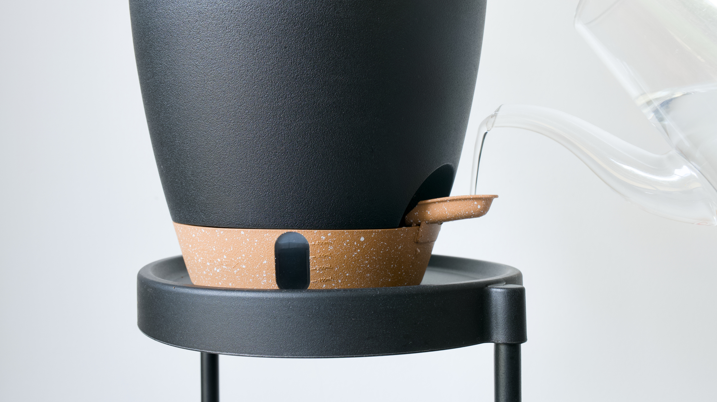 Water is poured from a glass pitcher into the reservoir of a modern black and terracotta self-watering planter.