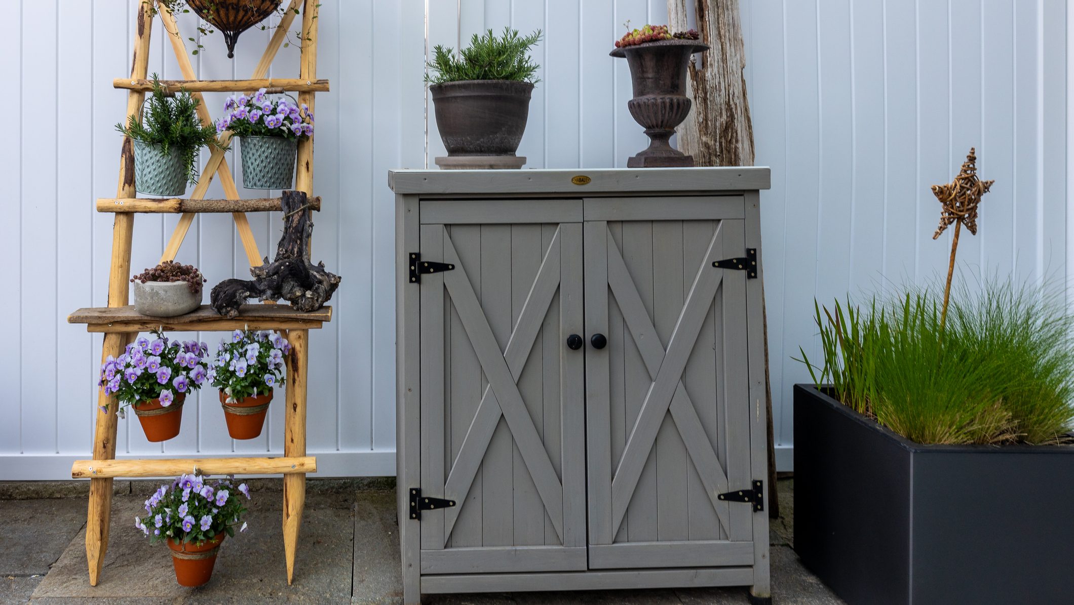 Gray garden cabinet positioned against a white fence, featuring potted plants like pansy and rosemary, decorative driftwood, and a wicker star, creating a charming outdoor space with rustic and modern elements
