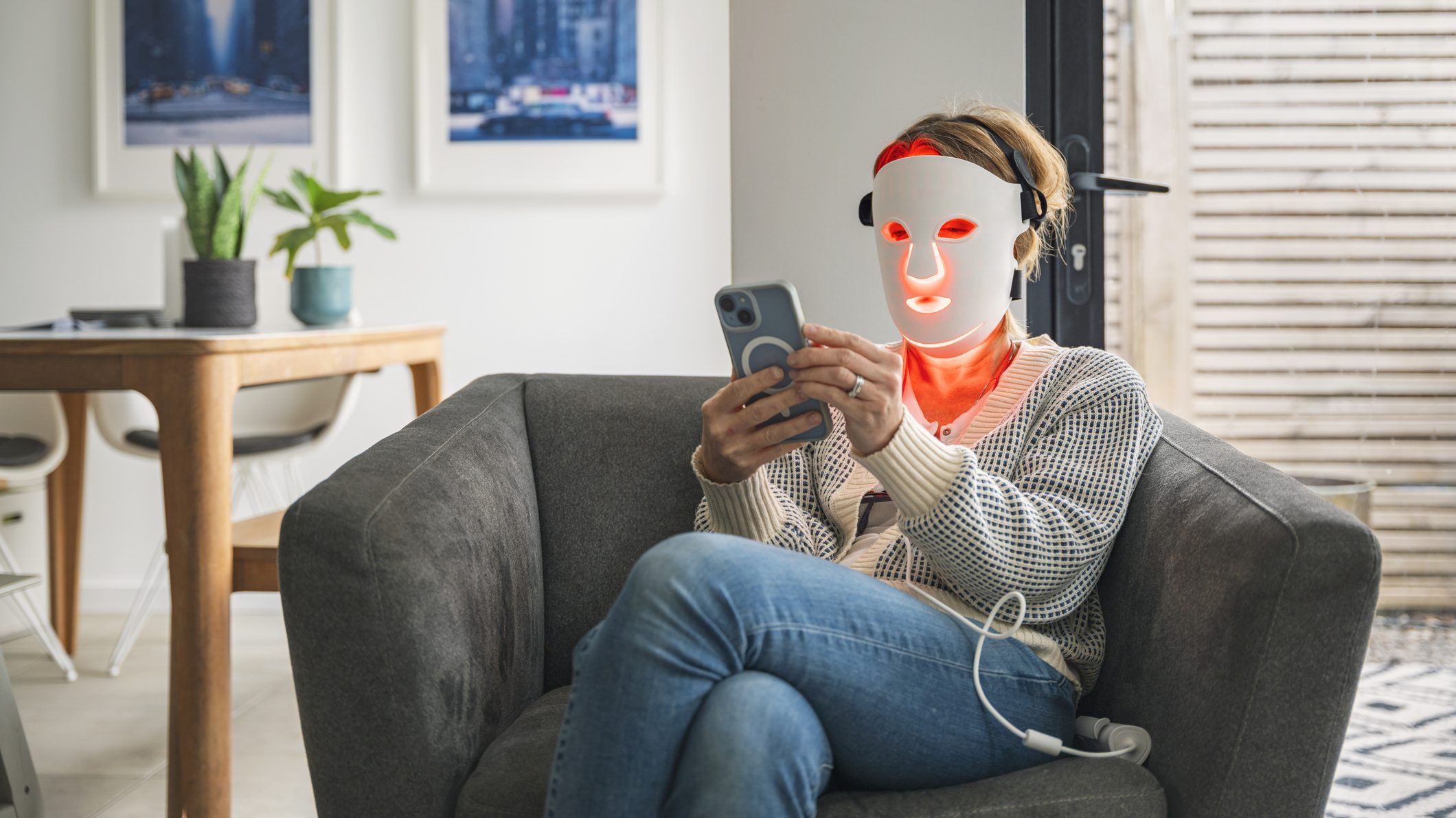 A mature woman uses her red LED face mask (red light therapy) whilst relaxing at home in an armchair. She uses her smartphone