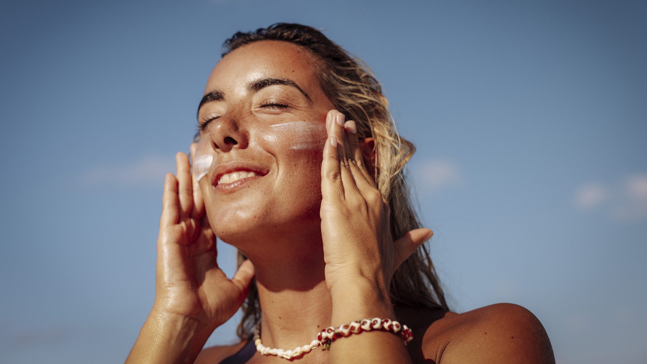 A happy young woman with sun-kissed skin applies sun cream to her face under a bright blue sky on a sunny beach in the Maldives