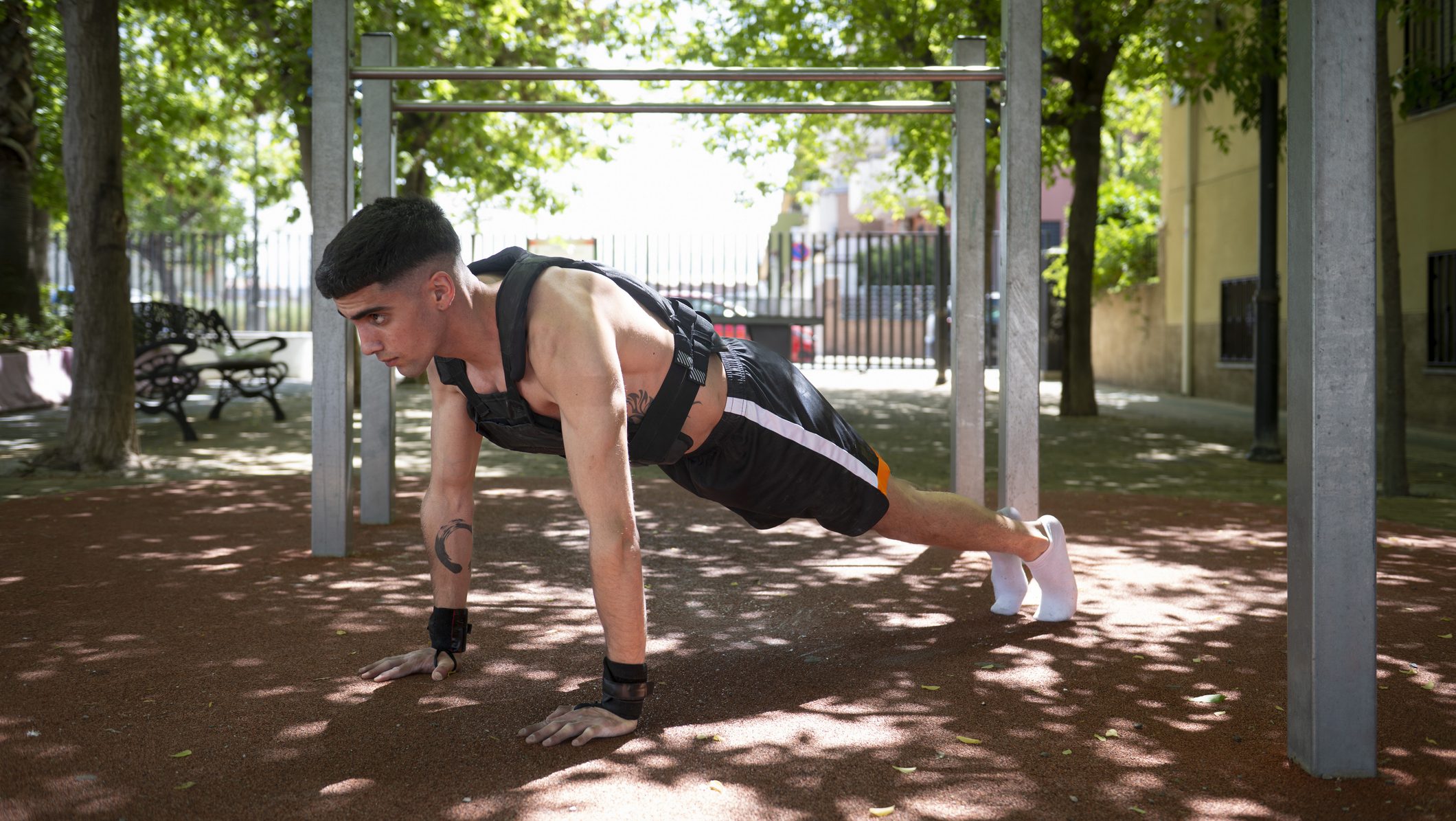 Determined male athlete performing plank exercise, part of calisthenics workout, wearing weighted vest in outdoor park setting, demonstrating strength and fitness