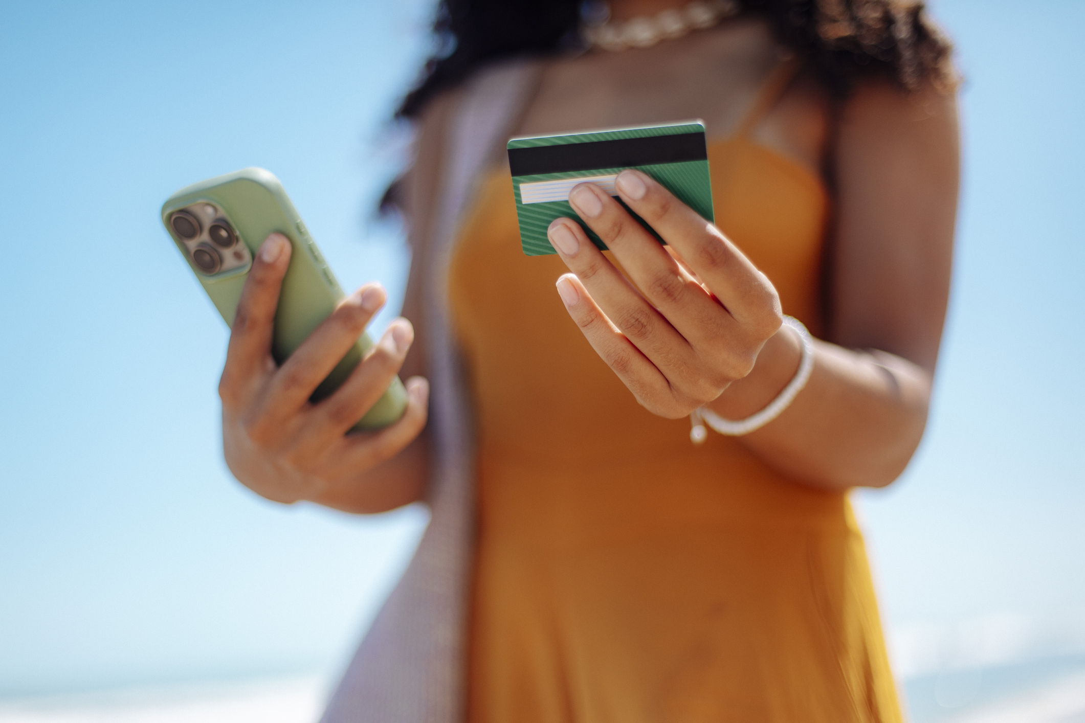 Close-up shot of a woman holding a smartphone and a credit card while standing on a sunny beach. The scene captures the concept of mobile banking, online shopping, digital payments, and modern lifestyle on vacation. Soft focus on the hands and card creates a bright, summery atmosphere with blue sky and ocean in the background. Ideal for illustrating fintech, travel purchases, wireless transactions, and summer leisure.