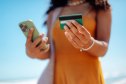 Close-up shot of a woman holding a smartphone and a credit card while standing on a sunny beach. The scene captures the concept of mobile banking, online shopping, digital payments, and modern lifestyle on vacation. Soft focus on the hands and card creates a bright, summery atmosphere with blue sky and ocean in the background. Ideal for illustrating fintech, travel purchases, wireless transactions, and summer leisure.