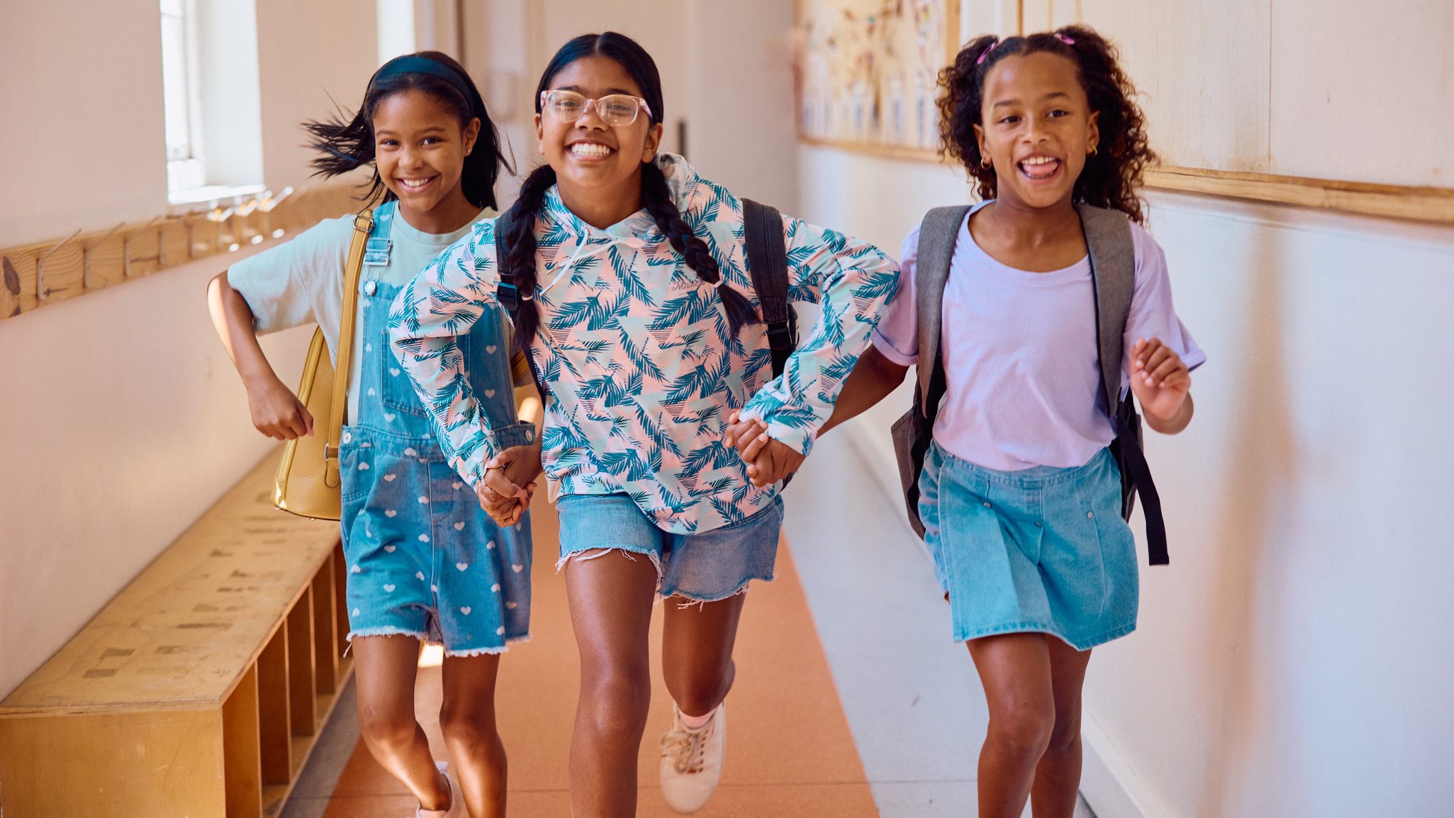 Three elementary school girls running together through a bright school corridor with backpacks while laughing and enjoying a playful moment between classes.