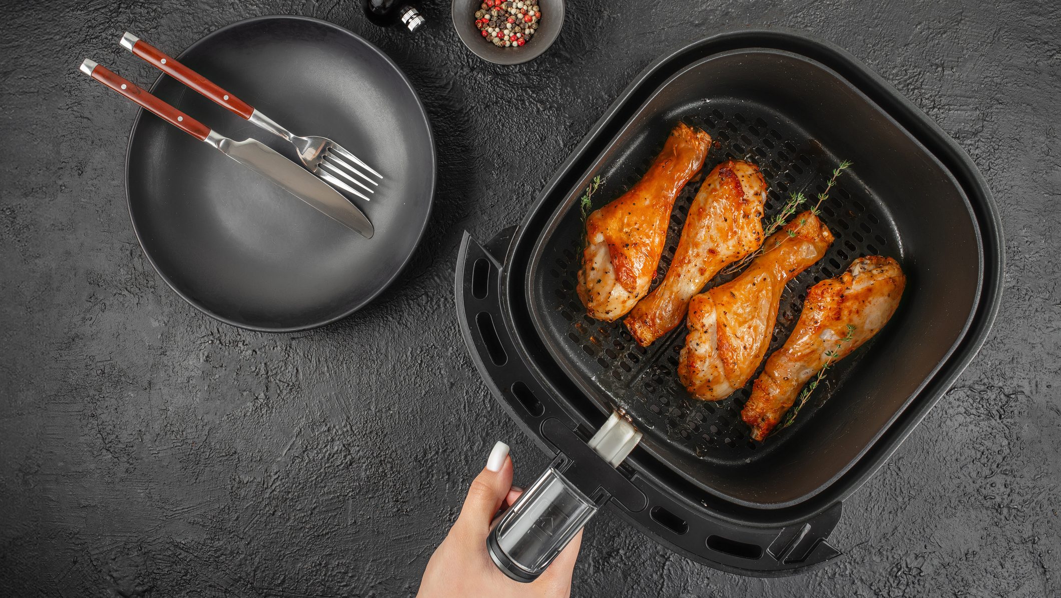 Woman holding air fryer basket with golden chicken drumsticks and herbs on black background. Top view healthy cooking concept.