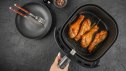 Woman holding air fryer basket with golden chicken drumsticks and herbs on black background. Top view healthy cooking concept.
