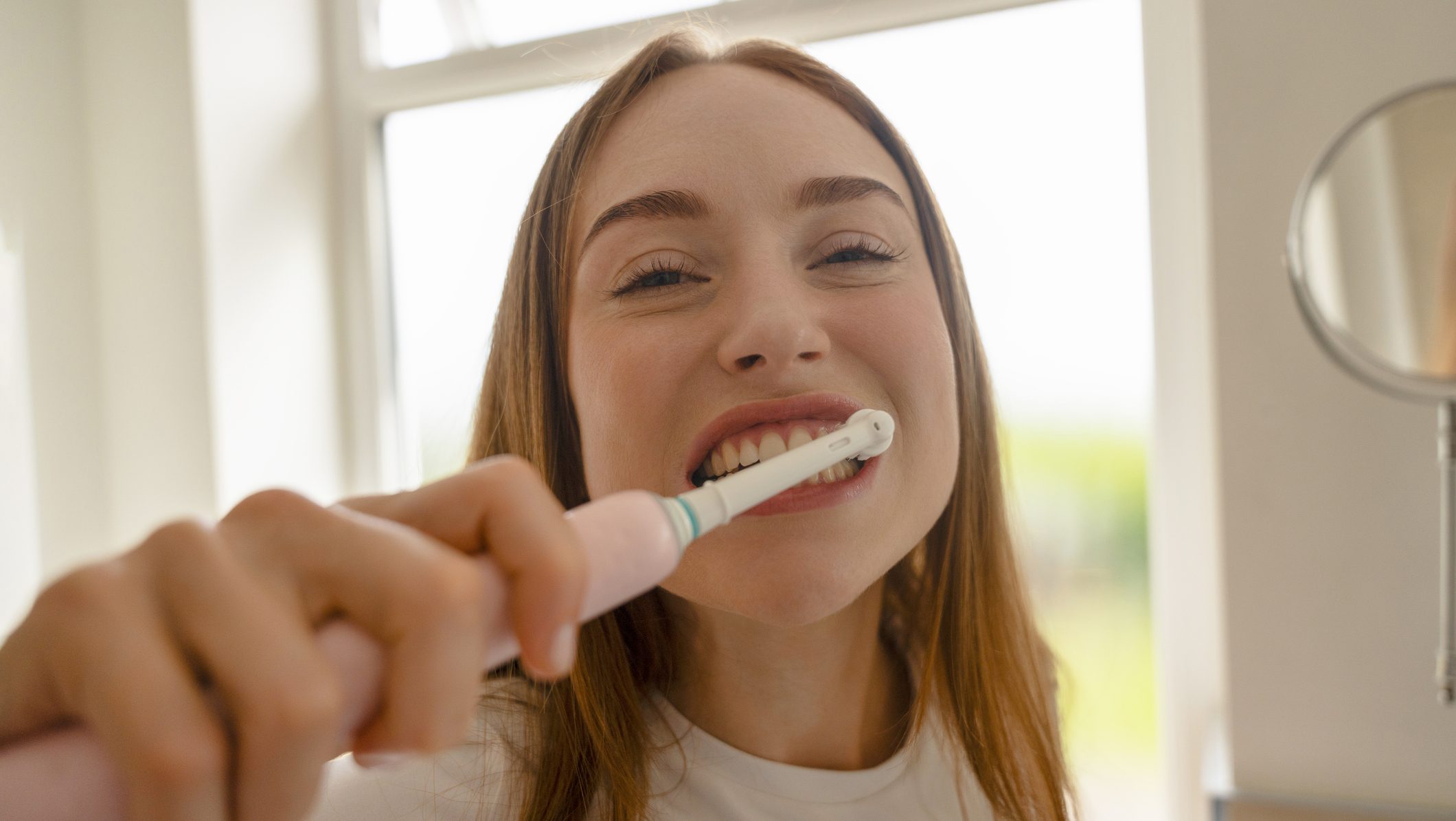 A young woman stands in a bathroom near a window. She holds an electric toothbrush and smiles while brushing her teeth. Morning light fills the space.