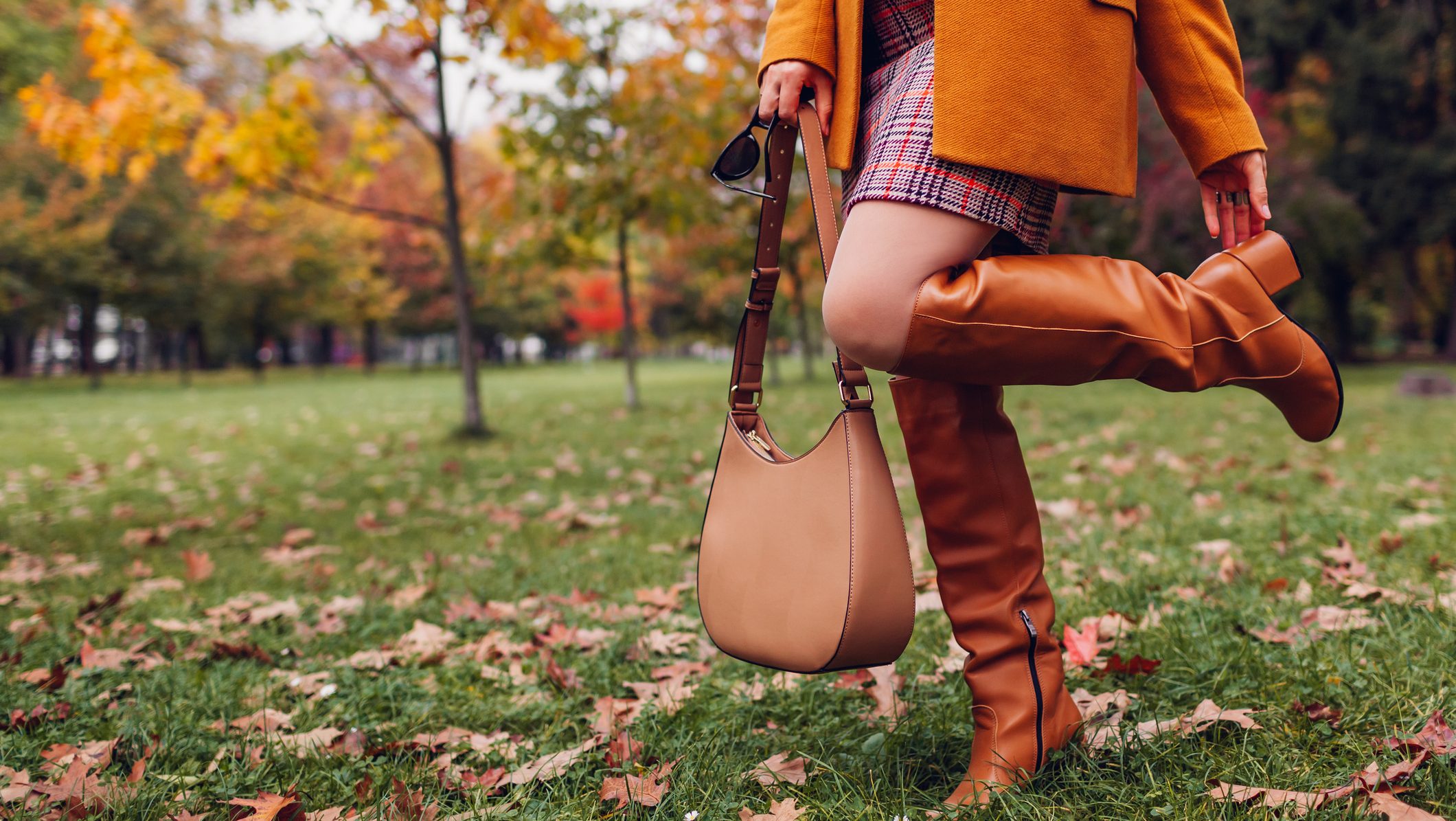 Orange knee high boots. Fashionable woman wearing stylish blazer plaid mini skirt walking in fall park with beige handbag among leaves. Copy space