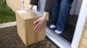 A medium unrecognisable shot of a woman bending down to pick up a package left at her doorstep. The scene highlights the convenience of home delivery, online shopping, and contactless drop-offs.
