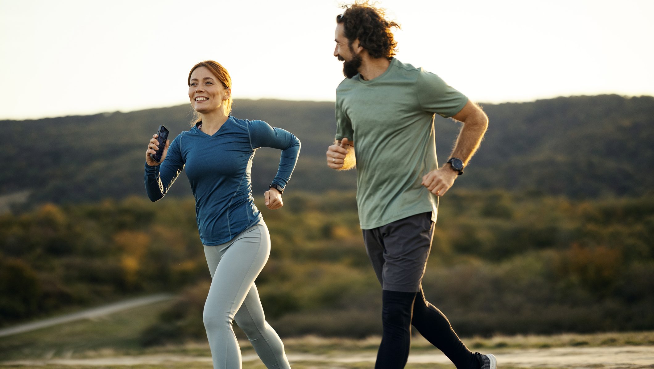 Happy couple running outdoors at sunset, smiling during a workout in a natural park for health and fitness