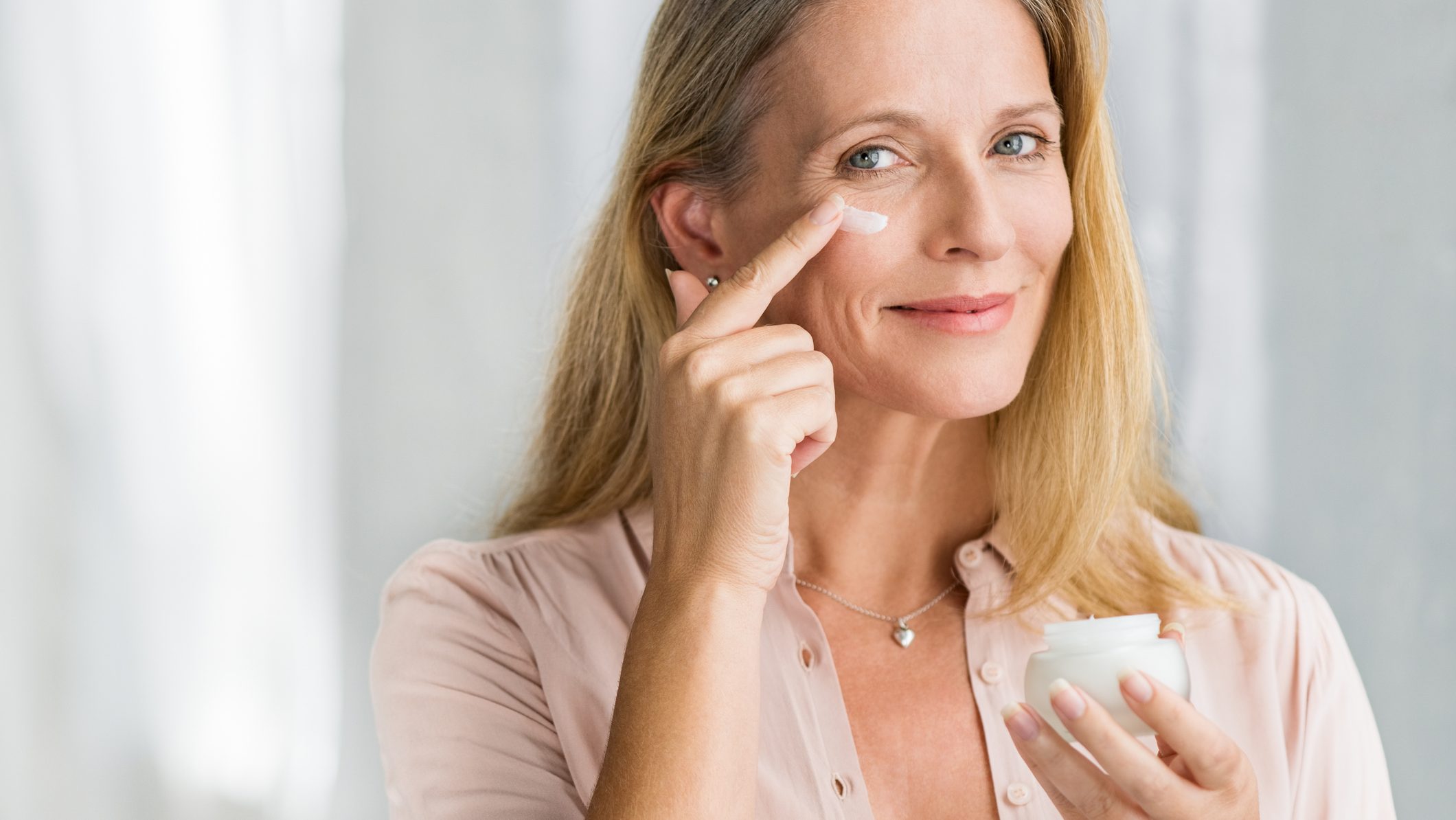 Smiling senior woman applying anti-aging lotion to remove dark circles under eyes. Happy mature woman using cosmetic cream to hide wrinkles below eyes. Lady using day moisturizer to counteract the aging of the skin.