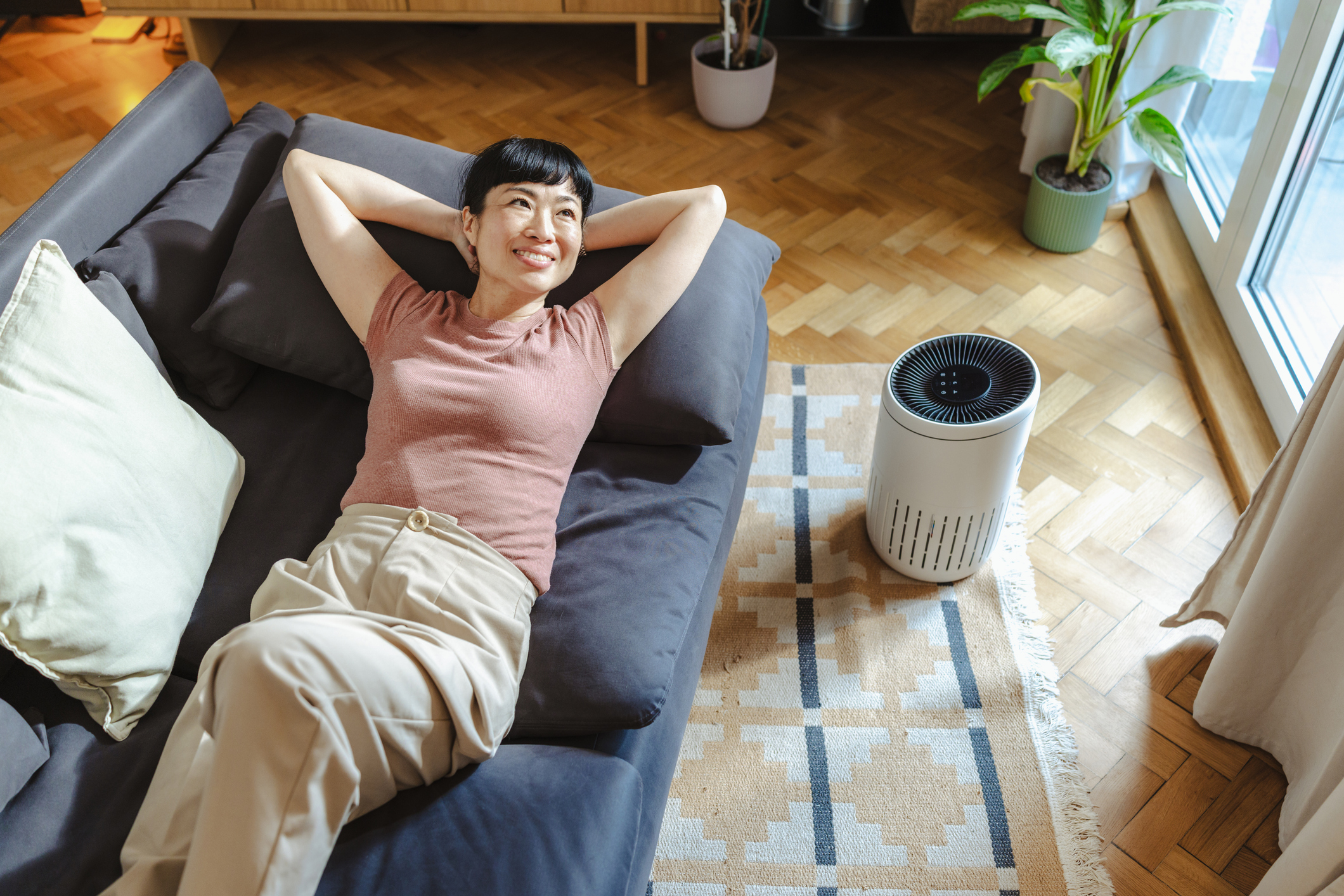 A relaxed woman lies on a sofa in a bright living room, smiling with hands behind her head beside an air purifier. Warm sunlight, indoor plants and a calm, comfortable atmosphere convey wellbeing and clean air.