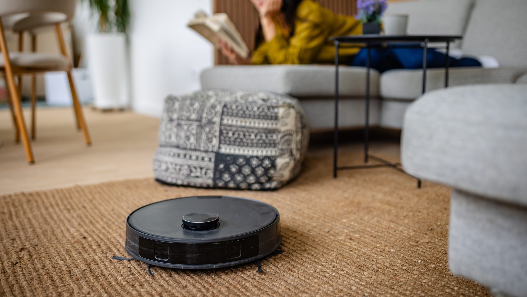 A woman lounges on a sofa, reading a book as a robotic vacuum operates on the floor. The cozy living room features a stylish rug, decorative pillows, and a potted plant, creating a modern, relaxed atmosphere.